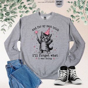 May include: A sport grey crewneck sweatshirt featuring a black and white illustration of a kitten wearing a pink party hat and holding a star-tipped wand. The text reads: and for my next trick i'll forget what i was doing, surrounded by stars.