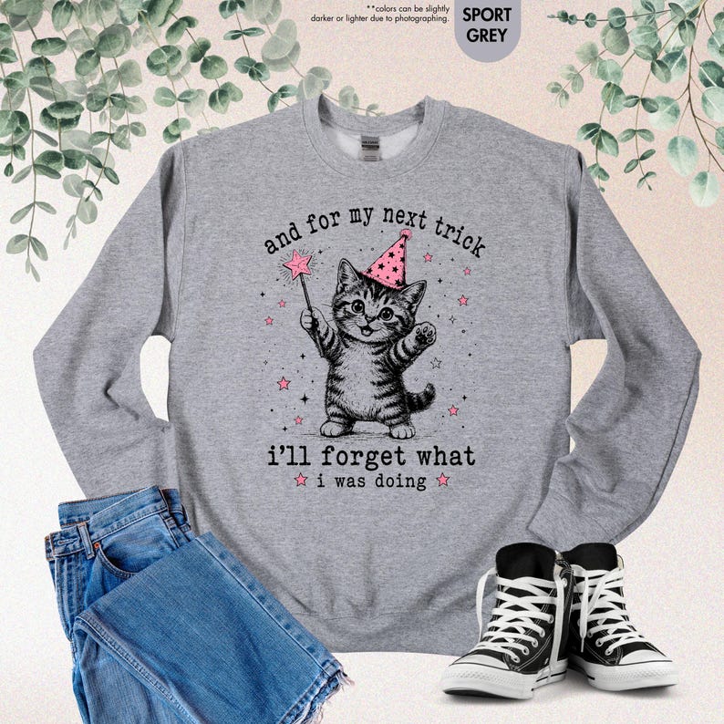 May include: A sport grey crewneck sweatshirt featuring a black and white illustration of a kitten wearing a pink party hat and holding a star-tipped wand. The text reads: and for my next trick i'll forget what i was doing, surrounded by stars.