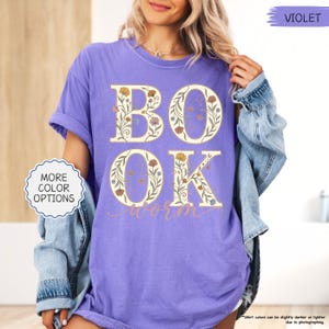 以下が含まれることがあります： 紫色のTシャツ。文字「BOOK WORM」が花柄のデザインで描かれています。文字はベージュで、様々な色の花のアクセントが付いています。ゆったりとしたフィット感と半袖です。デニムジャケットが肩にかけられています。