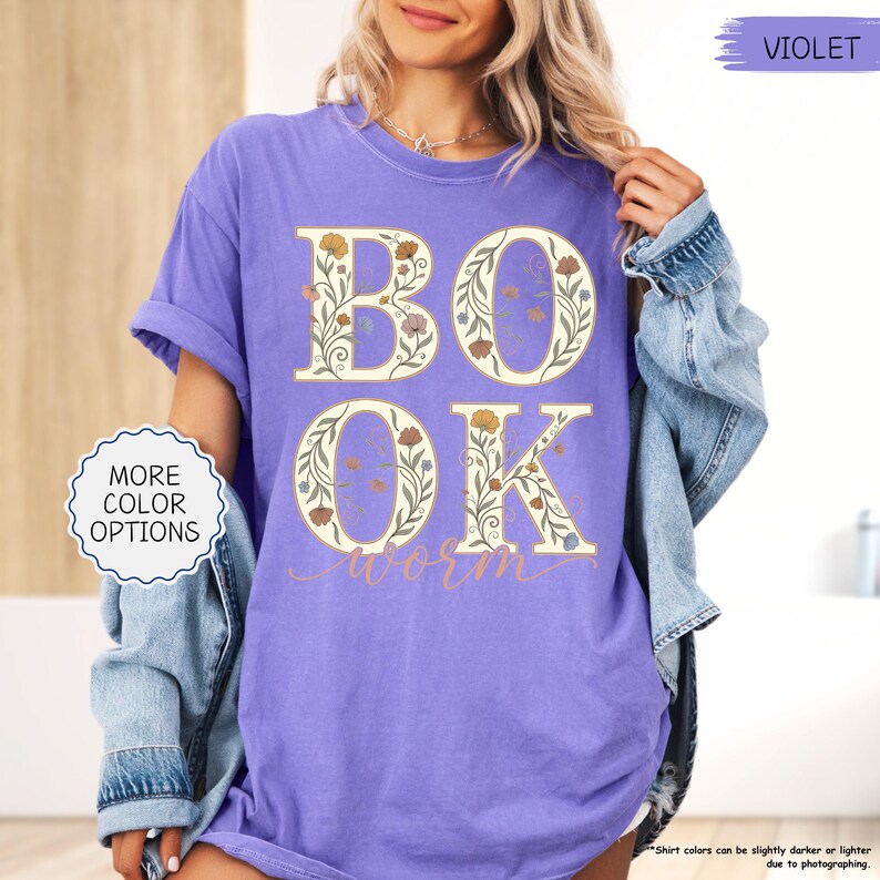 以下が含まれることがあります： 紫色のTシャツ。文字「BOOK WORM」が花柄のデザインで描かれています。文字はベージュで、様々な色の花のアクセントが付いています。ゆったりとしたフィット感と半袖です。デニムジャケットが肩にかけられています。