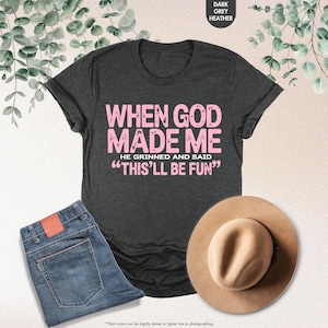 May include: Dark grey heather t-shirt with the text "WHEN GOD MADE ME HE GRINNED AND SAID 'THIS'LL BE FUN'" in pink. The image also includes blue jeans and a tan hat. The shirt is a casual wear item.