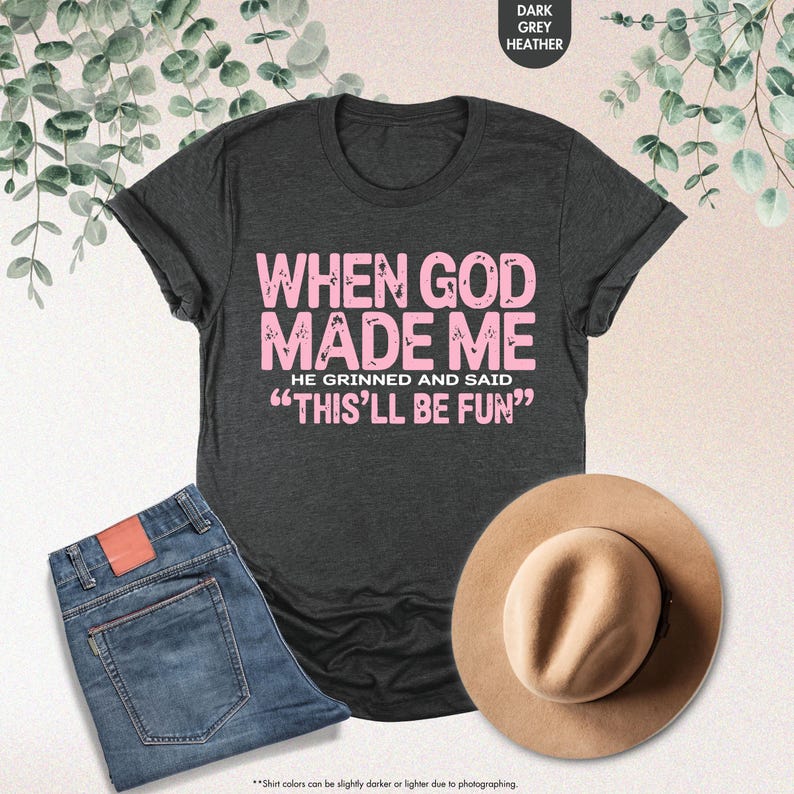 May include: Dark grey heather t-shirt with the text "WHEN GOD MADE ME HE GRINNED AND SAID 'THIS'LL BE FUN'" in pink. The image also includes blue jeans and a tan hat. The shirt is a casual wear item.