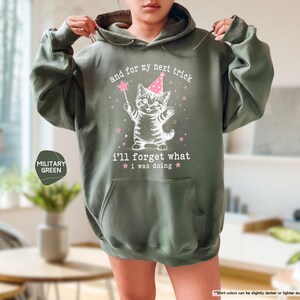 May include: A military green hooded sweatshirt featuring a white graphic of a kitten wearing a pink party hat and holding a magic wand. The text reads: and for my next trick i'll forget what i was doing, surrounded by small pink stars.