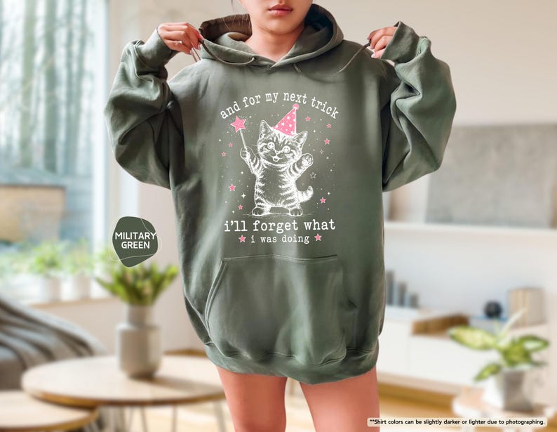 May include: A military green hooded sweatshirt featuring a white graphic of a kitten wearing a pink party hat and holding a magic wand. The text reads: and for my next trick i'll forget what i was doing, surrounded by small pink stars.