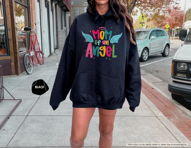 May include: Black hooded sweatshirt with the text "MOM of an Angel" in colorful letters and angel wings. The sweatshirt has a front pocket and a drawstring hood. The word "BLACK" is in a black circle.