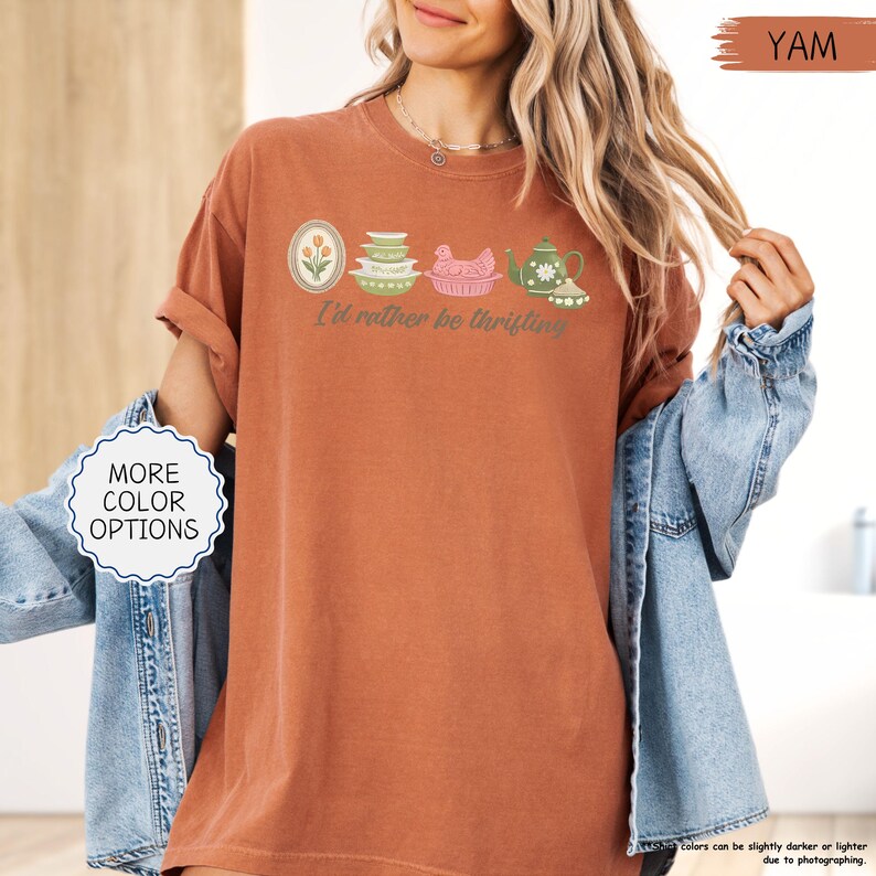 Pu&ograve; includere: T-shirt color ruggine con la scritta "I'd rather be thrifting" e una grafica di oggetti da cucina vintage. La grafica include un motivo floreale, ciotole impilate, un pollo e una teiera. La maglietta ha le maniche arrotolate.