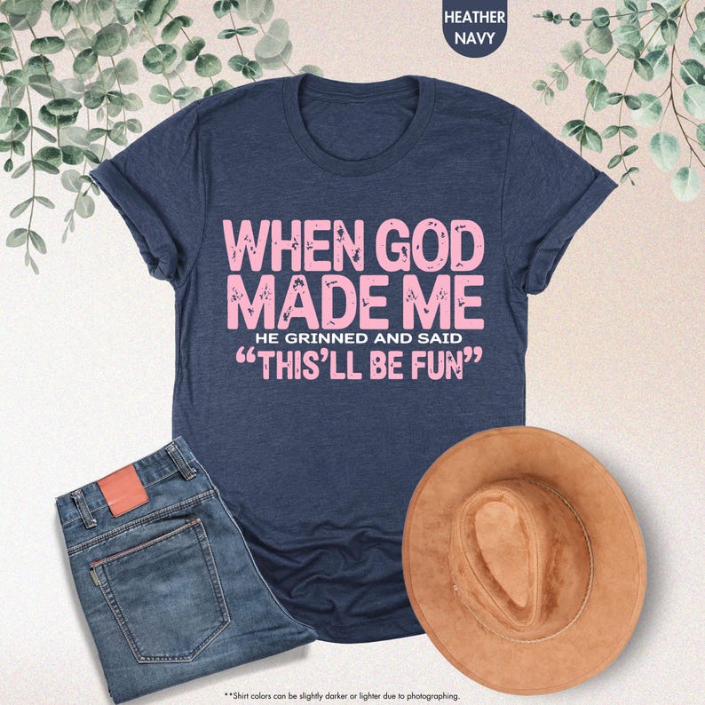 May include: Heather navy t-shirt with the text "WHEN GOD MADE ME HE GRINNED AND SAID 'THIS'LL BE FUN'" in pink. The image also includes a pair of blue jeans and a brown hat.