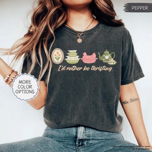Pu&ograve; includere: T-shirt grigio scuro con la frase "I'd rather be thrifting" e illustrazioni di utensili da cucina vintage. Il design include un tulipano incorniciato, ciotole impilate, una gallina in un cestino e una teiera.