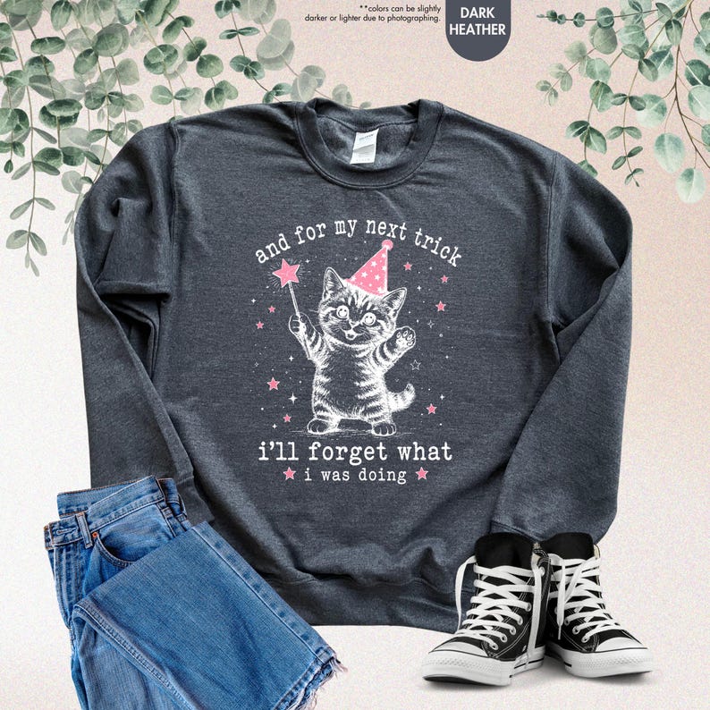 May include: A dark heather gray crewneck sweatshirt featuring a white graphic of a kitten wearing a pink party hat and holding a star wand. The text reads, and for my next trick i'll forget what i was doing, surrounded by small pink stars.