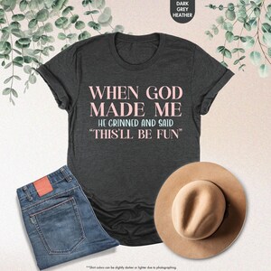 May include: Dark grey heather t-shirt with the text "WHEN GOD MADE ME HE GRINNED AND SAID 'THIS'LL BE FUN'" in pink and blue. The image also shows blue jeans and a tan hat.