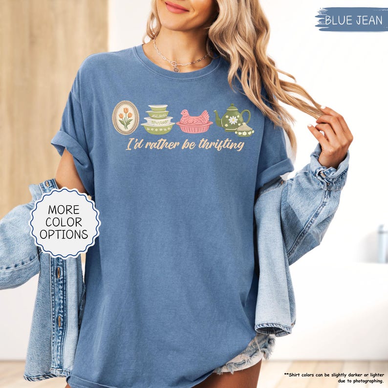 Pu&ograve; includere: T-shirt blu jeans con la frase "I'd rather be thrifting" e una grafica di utensili da cucina vintage. La grafica include un ovale floreale, ciotole impilate, una gallina rosa e una teiera. "More color options" &egrave; in un cerchio bianco.