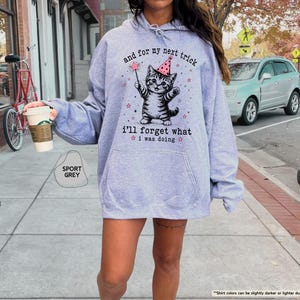 May include: A sport grey hooded sweatshirt featuring a graphic of a kitten wearing a pink polka-dot party hat and holding a magic wand. The text reads, "and for my next trick i'll forget what i was doing," surrounded by small pink stars.