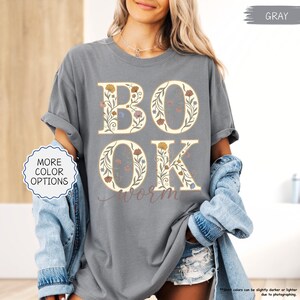 以下が含まれることがあります： 大きな花柄の文字で「BOOK」と「OK」と書かれたグレーのTシャツ。「worm」の文字が筆記体で下に書かれています。シャツには「MORE COLOR OPTIONS」ボタンも付いています。