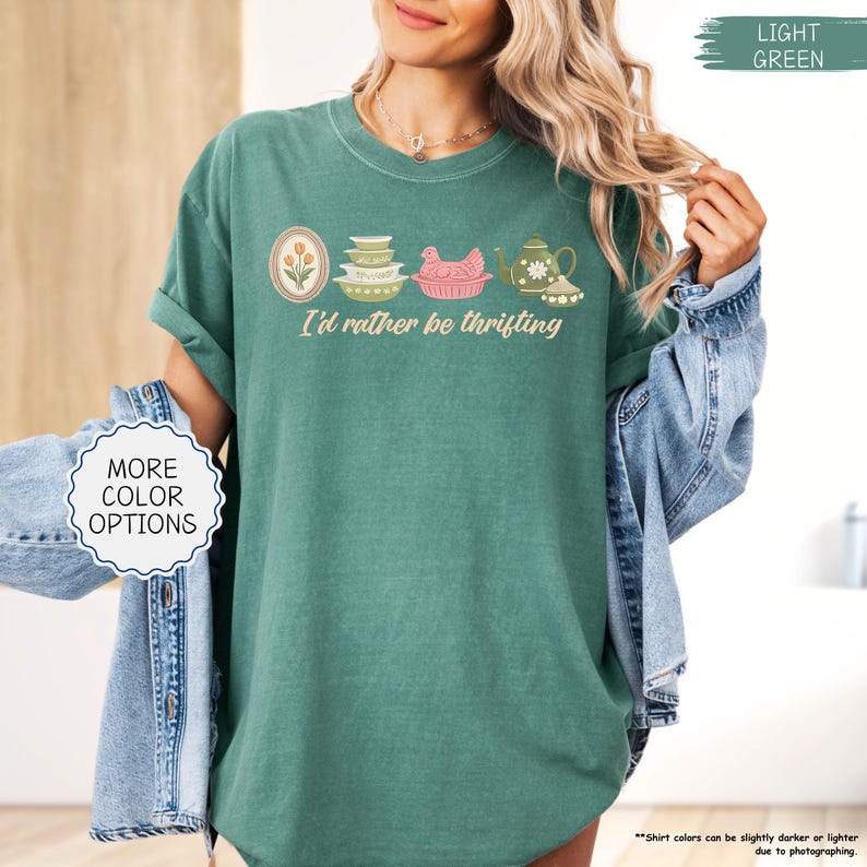 Pu&ograve; includere: T-shirt verde chiaro con la scritta "I'd rather be thrifting" e una grafica di oggetti da cucina vintage. La grafica include un design floreale, ciotole, un piatto a forma di pollo e una teiera. La maglietta ha le maniche arrotolate.