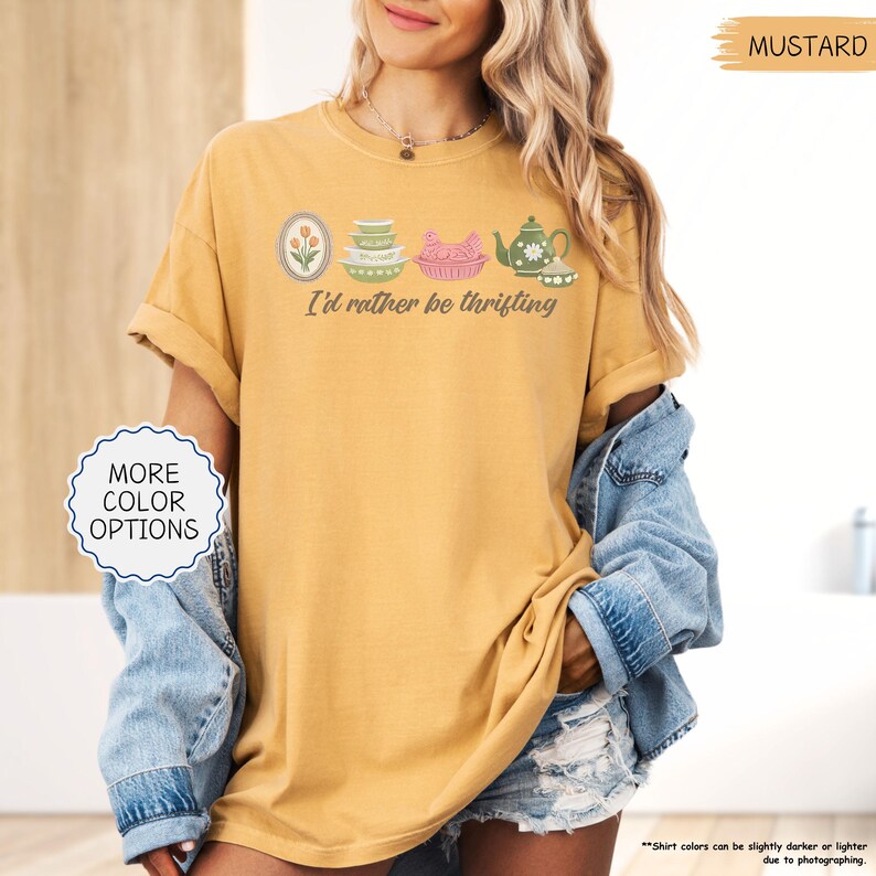 Pu&ograve; includere: T-shirt color senape con la frase "I'd rather be thrifting" e una grafica di utensili da cucina vintage. La grafica include un'immagine con cornice floreale, piatti impilati, un piatto rosa a forma di pollo e una teiera.