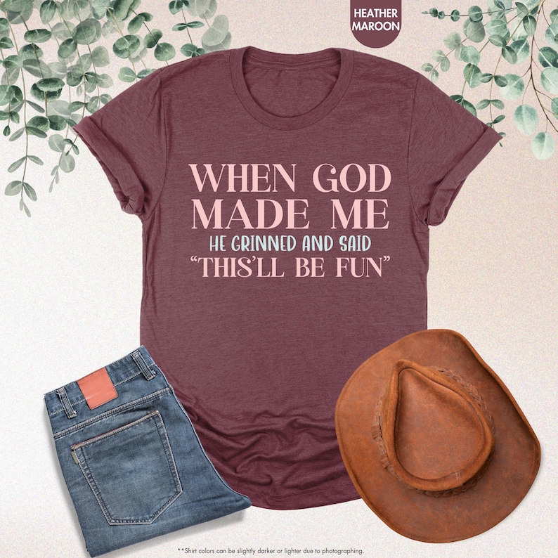 May include: Heather maroon t-shirt with the text "WHEN GOD MADE ME HE GRINNED AND SAID 'THIS'LL BE FUN'". The image also includes a pair of blue jeans and a brown cowboy hat.