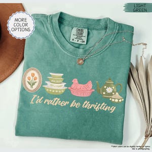 Pu&ograve; includere: T-shirt Comfort Colors verde chiaro con la scritta "I'd rather be thrifting" e una grafica di un fiore incorniciato, ciotole, una gallina, una teiera e una zuccheriera. Una collana d'argento &egrave; drappeggiata sulla maglietta.