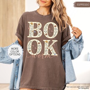 以下が含まれることがあります： フローラルデザインの「BOOK WORM」の文字が入ったエスプレッソカラーのTシャツ。文字はクリーム色で縁取られ、カラフルな花で埋められています。シャツは袖をまくり上げ、デニムジャケットと合わせています。