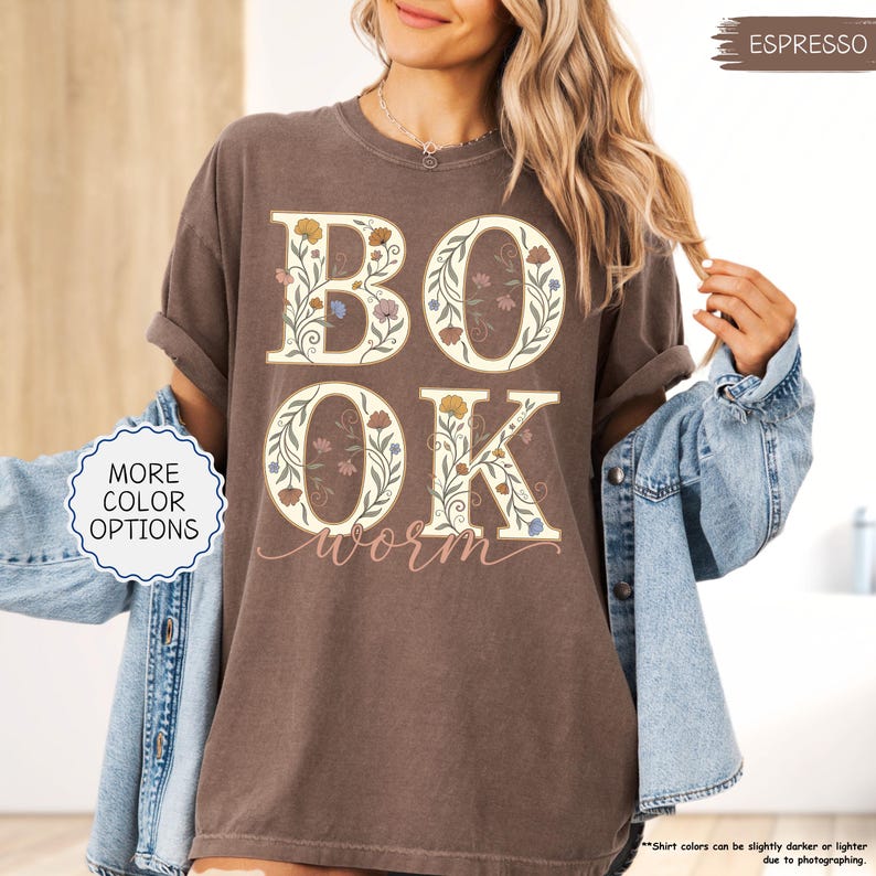 以下が含まれることがあります： フローラルデザインの「BOOK WORM」の文字が入ったエスプレッソカラーのTシャツ。文字はクリーム色で縁取られ、カラフルな花で埋められています。シャツは袖をまくり上げ、デニムジャケットと合わせています。