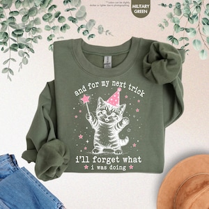 May include: A military green crewneck sweatshirt featuring a white graphic of a kitten wearing a pink party hat and holding a star-tipped wand. The text reads, and for my next trick i'll forget what i was doing, surrounded by small pink stars.