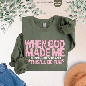 May include: Military green sweatshirt with the text "WHEN GOD MADE ME HE GRINNED AND SAID 'THIS'LL BE FUN'" in pink. The sweatshirt is a crew neck style and has long sleeves. The background includes denim jeans and a hat.