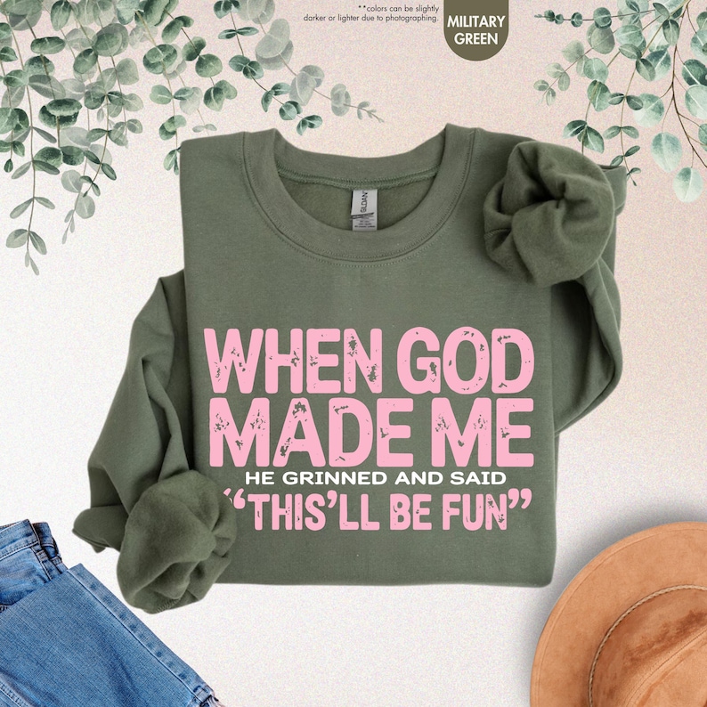 May include: Military green sweatshirt with the text "WHEN GOD MADE ME HE GRINNED AND SAID 'THIS'LL BE FUN'" in pink. The sweatshirt is a crew neck style and has long sleeves. The background includes denim jeans and a hat.