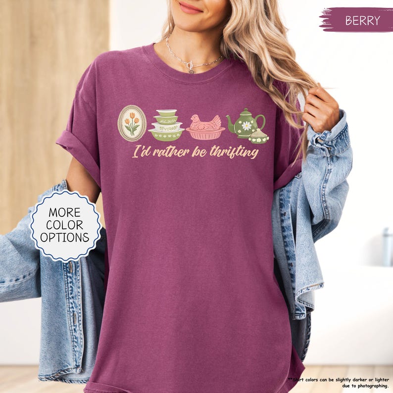 Pu&ograve; includere: T-shirt color bacca con la frase "I'd rather be thrifting" e una grafica di utensili da cucina vintage. Il design include un ovale floreale, ciotole impilate, una gallina in un cestino e una teiera. La maglietta ha le maniche arrotolate.