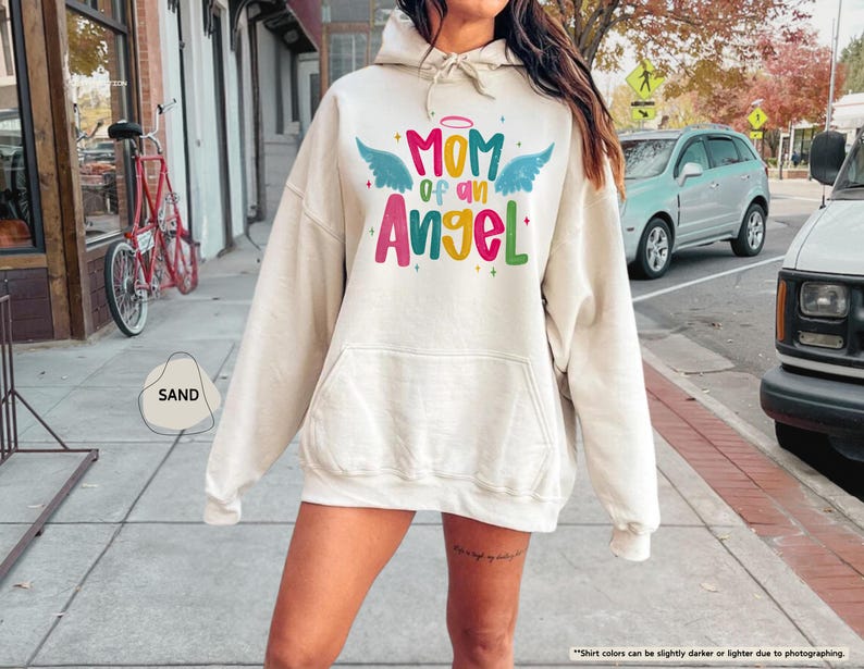 May include: A sand-colored hoodie with a front pocket and drawstring hood. The front features the text "MOM of an Angel" in colorful lettering with angel wings and a halo. The hoodie is a casual style.