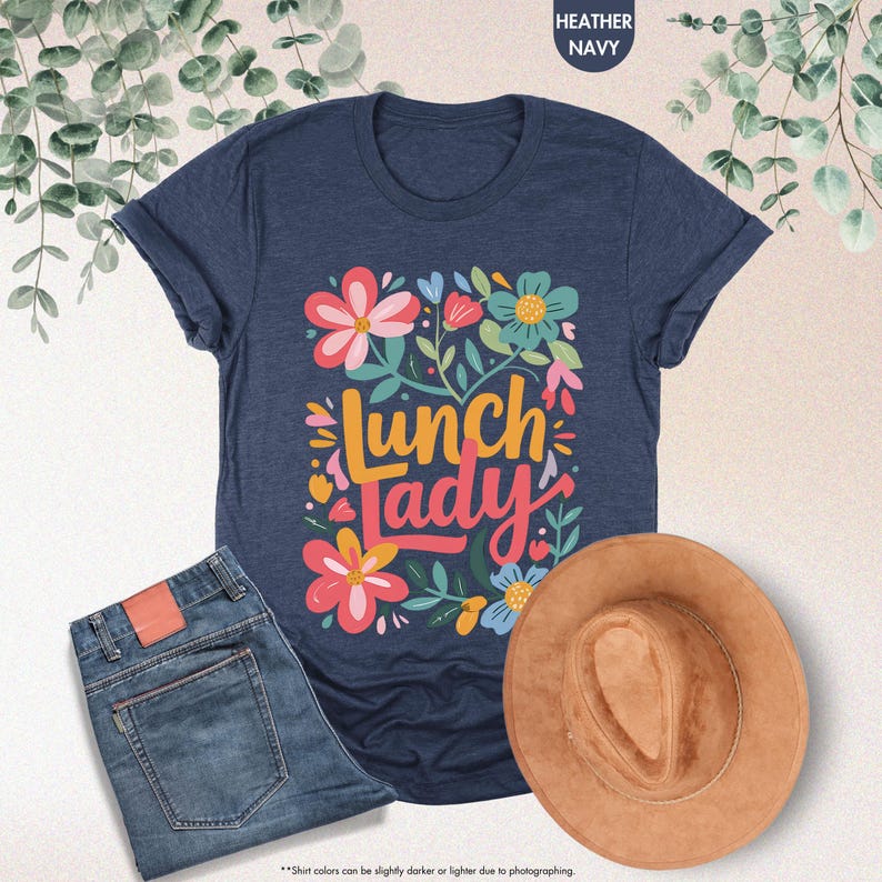 Pode incluir: T-shirt azul marinho com design floral e a frase "Lunch Lady". A imagem tamb&eacute;m inclui um par de jeans azuis e um chap&eacute;u castanho. A t-shirt &eacute; feita de um material macio.