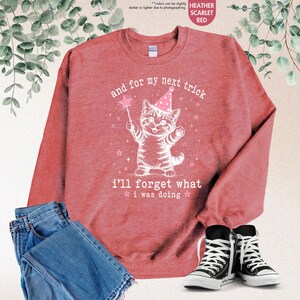 May include: A heather scarlet red crewneck sweatshirt featuring a white line art illustration of a kitten wearing a party hat and holding a star wand. The text reads: and for my next trick i'll forget what i was doing. Blue jeans and black sneakers.