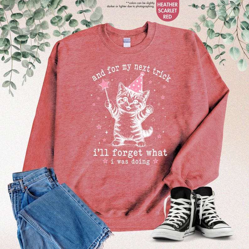 May include: A heather scarlet red crewneck sweatshirt featuring a white line art illustration of a kitten wearing a party hat and holding a star wand. The text reads: and for my next trick i'll forget what i was doing. Blue jeans and black sneakers.