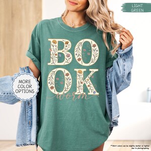 以下が含まれることがあります： 大きな花柄の文字で「BOOK」と「OK」と書かれた、明るい緑色のTシャツ。「worm」の文字が筆記体で下に書かれています。シャツの袖はロールアップされており、「MORE COLOR OPTIONS」の文字が見えます。