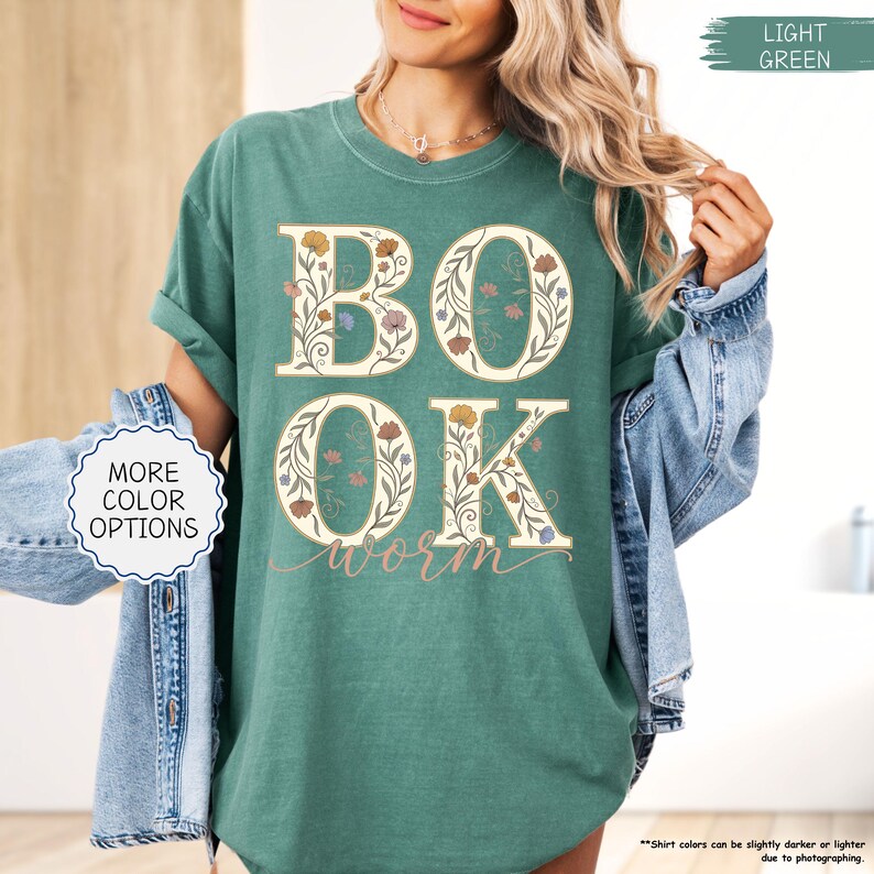 以下が含まれることがあります： 大きな花柄の文字で「BOOK」と「OK」と書かれた、明るい緑色のTシャツ。「worm」の文字が筆記体で下に書かれています。シャツの袖はロールアップされており、「MORE COLOR OPTIONS」の文字が見えます。