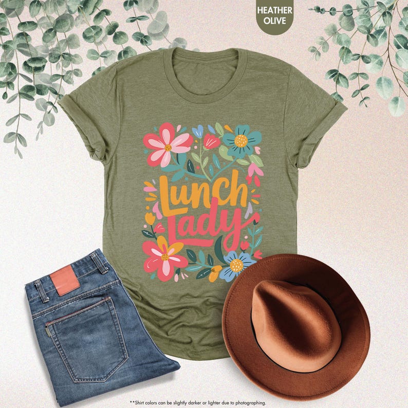 Pode incluir: T-shirt verde azeitona mesclado com o texto "Lunch Lady" em um design floral colorido. A imagem tamb&eacute;m inclui um par de jeans azuis e um chap&eacute;u marrom.