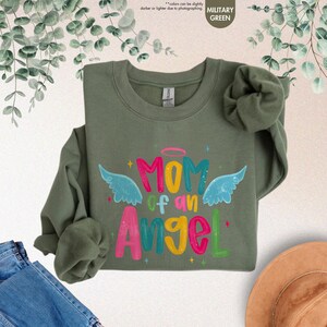 May include: Military green sweatshirt with the text "MOM of an Angel" in colorful letters, angel wings, and a halo. The sweatshirt is a crew neck style, and the sleeves are folded. The background includes denim jeans and a hat.