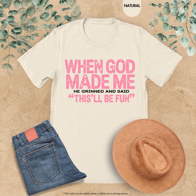 May include: Cream-colored t-shirt with the text "WHEN GOD MADE ME HE GRINNED AND SAID 'THIS'LL BE FUN'" in pink. The shirt is paired with blue jeans and a brown hat. The image is set against a neutral background.