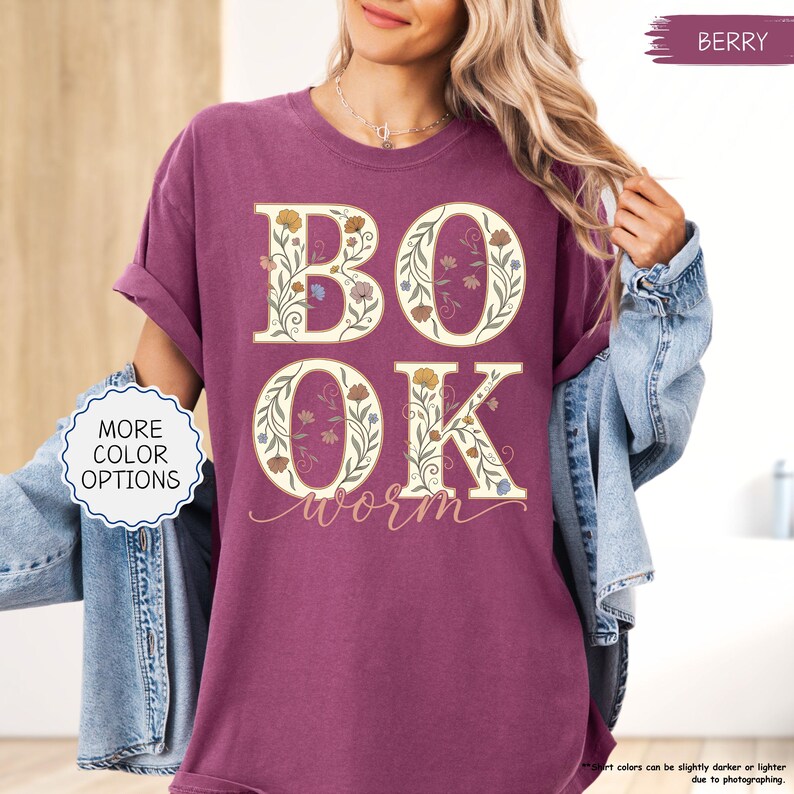 以下が含まれることがあります： ベリー色のTシャツ。花柄のデザインで「BOOK WORM」の文字が描かれています。文字はクリーム色で縁取られ、黄色、青、オレンジ色の花で埋められています。シャツの袖はロールアップされています。