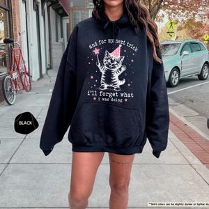 May include: A black hooded sweatshirt featuring a graphic of a kitten wearing a pink party hat and holding a star wand. The text reads, "and for my next trick i'll forget what i was doing," surrounded by small pink stars and white sparkles.