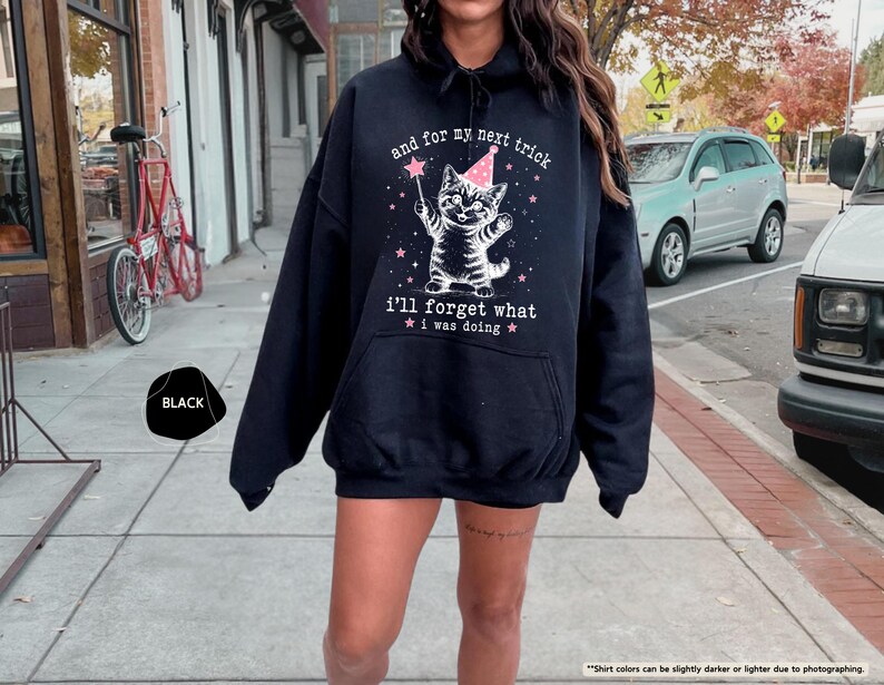 May include: A black hooded sweatshirt featuring a graphic of a kitten wearing a pink party hat and holding a star wand. The text reads, "and for my next trick i'll forget what i was doing," surrounded by small pink stars and white sparkles.