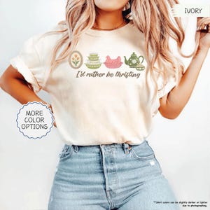 Pu&ograve; includere: T-shirt color avorio con la scritta "I'd rather be thrifting" e illustrazioni di un tulipano, ciotole, un pollo e una teiera. La modella indossa jeans blu. Sono disponibili opzioni di colore aggiuntive.