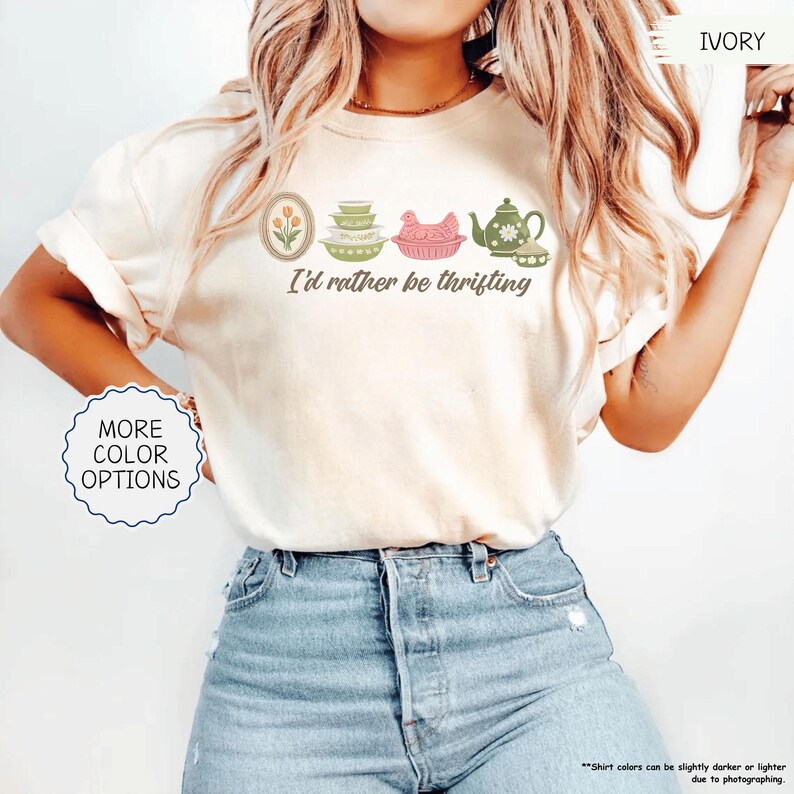 Pu&ograve; includere: T-shirt color avorio con la scritta "I'd rather be thrifting" e illustrazioni di un tulipano, ciotole, un pollo e una teiera. La modella indossa jeans blu. Sono disponibili opzioni di colore aggiuntive.