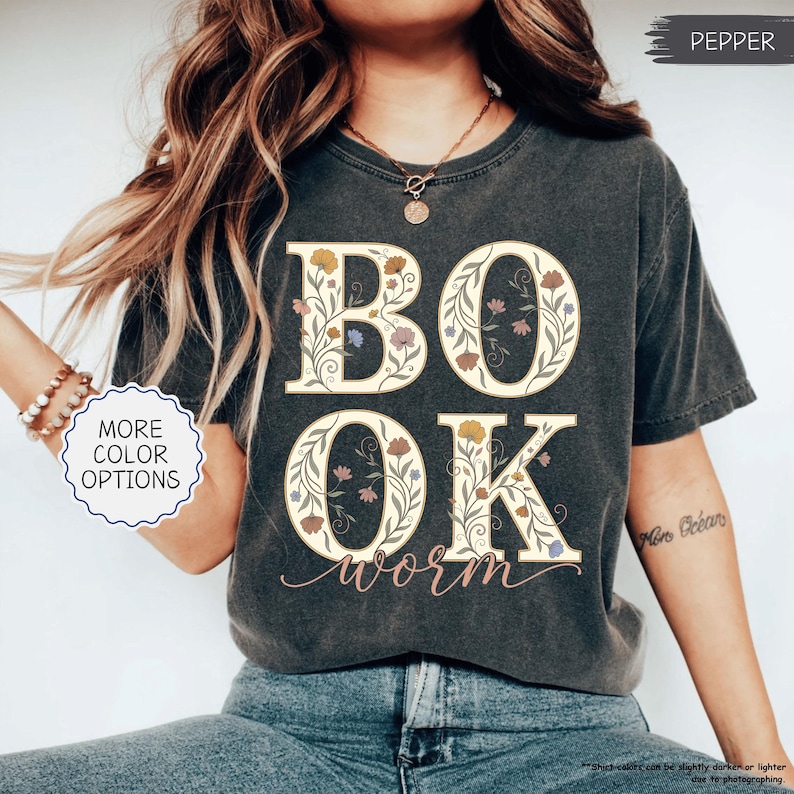 以下が含まれることがあります： 大きな花柄の文字で「BOOK」と「OK」と書かれた濃い灰色のTシャツ。「worm」の文字が筆記体で下に書かれています。シャツはヴィンテージ風で、色あせた外観です。「MORE COLOR OPTIONS」の文字が見えます。