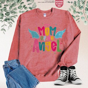May include: Heather scarlet red sweatshirt with the text "MOM of an Angel" in colorful letters and angel wings. The sweatshirt is paired with blue jeans and black and white sneakers.