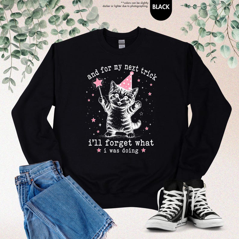 May include: A black long-sleeve sweatshirt featuring a white graphic of a kitten wearing a pink party hat and holding a star-tipped wand. The text reads, "and for my next trick i'll forget what i was doing," surrounded by small pink stars.