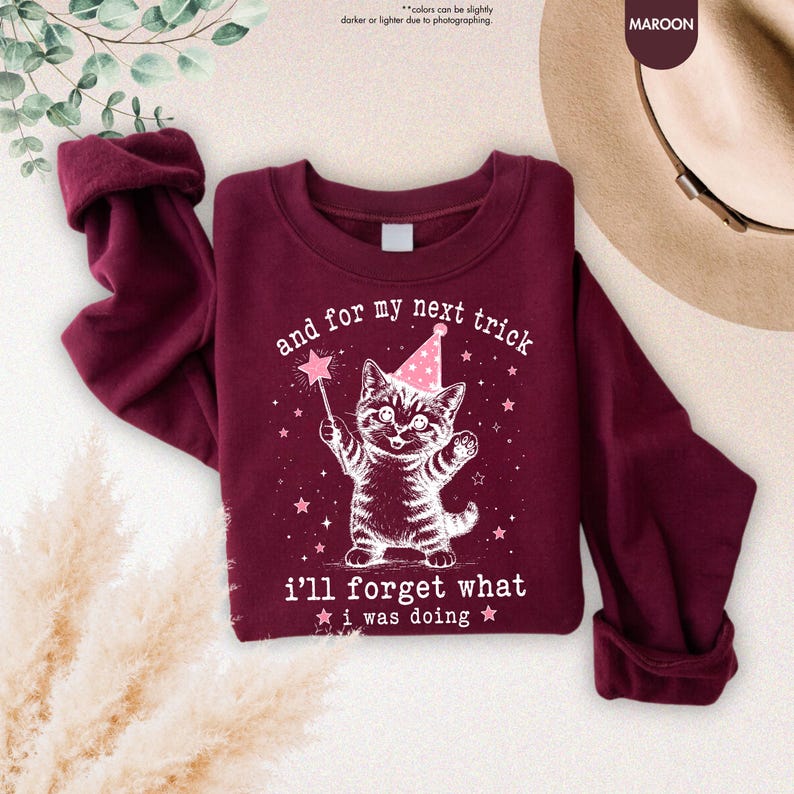 May include: A maroon long-sleeve sweatshirt featuring a white graphic of a kitten wearing a pink party hat and holding a star-tipped wand. The text reads, "and for my next trick i'll forget what i was doing," surrounded by small stars.