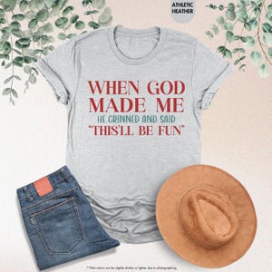 May include: A light gray t-shirt with the text "WHEN GOD MADE ME HE GRINNED AND SAID 'THIS'LL BE FUN'" in red and blue. The shirt is paired with blue jeans and a brown hat. The words "ATHLETIC HEATHER" are at the top.