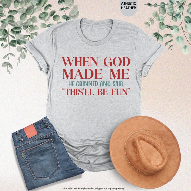 May include: A light gray t-shirt with the text "WHEN GOD MADE ME HE GRINNED AND SAID 'THIS'LL BE FUN'" in red and blue. The shirt is paired with blue jeans and a brown hat. The words "ATHLETIC HEATHER" are at the top.