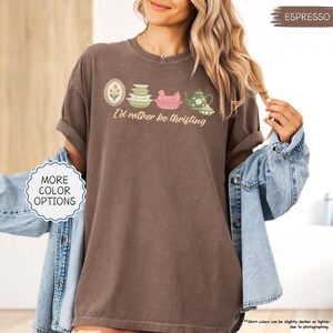 Pu&ograve; includere: T-shirt color espresso con la scritta "I'd rather be thrifting" e una grafica di utensili da cucina vintage. Il design include un ovale floreale, ciotole impilate, un pollo rosa e una teiera. La maglietta &egrave; indossata con una giacca di jeans.