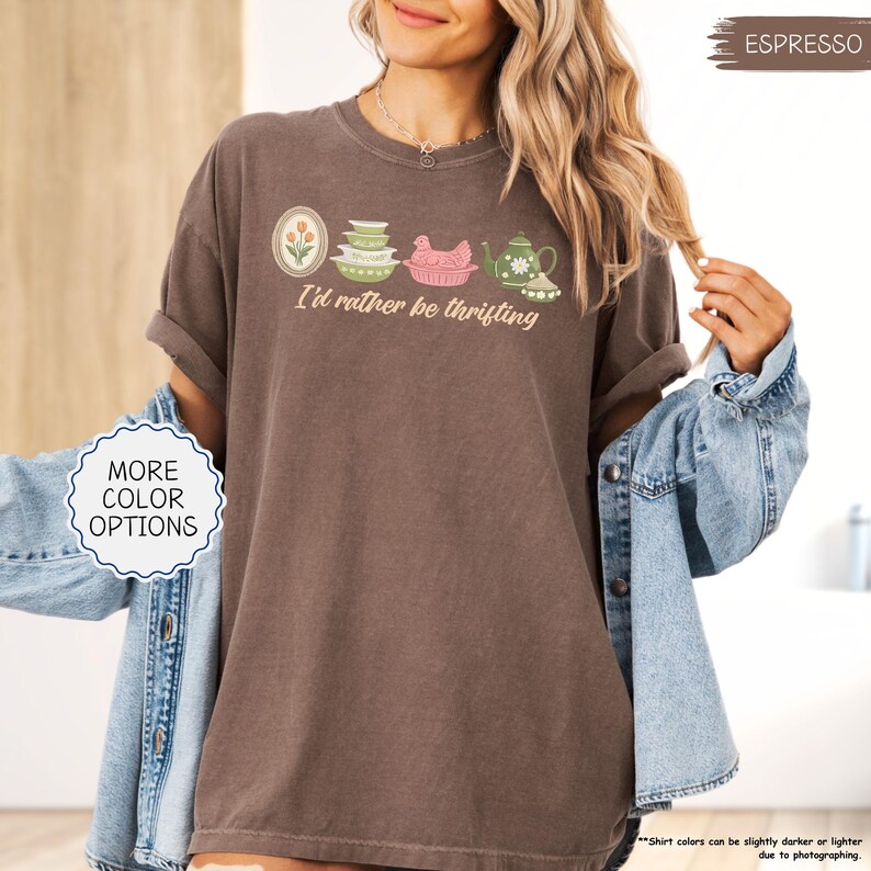 Pu&ograve; includere: T-shirt color espresso con la scritta "I'd rather be thrifting" e una grafica di utensili da cucina vintage. Il design include un ovale floreale, ciotole impilate, un pollo rosa e una teiera. La maglietta &egrave; indossata con una giacca di jeans.