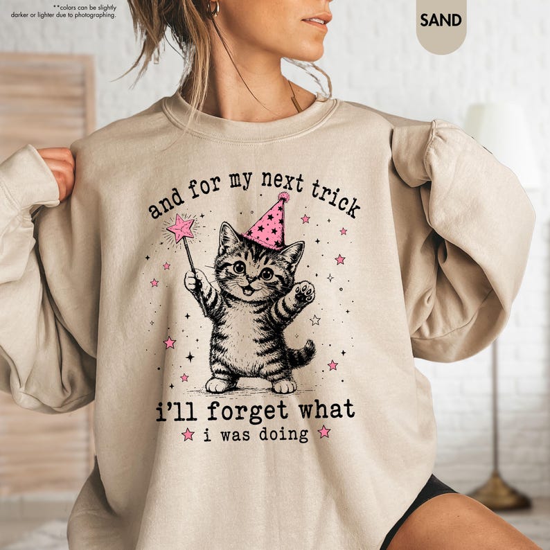 May include: A sand-colored crewneck sweatshirt featuring a graphic of a tabby kitten wearing a pink party hat, holding a star-tipped magic wand. The text reads, and for my next trick i'll forget what i was doing, surrounded by small pink stars.